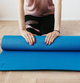 Female preparing for workout by rolling a blue yoga mat on wooden floor with dumbbells nearby.